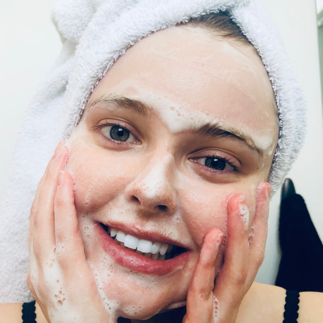 pretty girl smiling while washing her face with her hair in a towel and bubbly suds on her face 