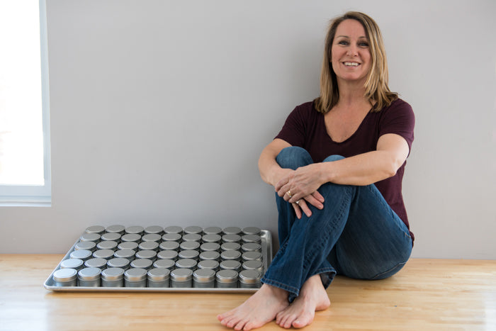 SiSi Georgian Bay founder Andrea Tranter sitting on the floor in jeans and a t-shirt and bare feet beside a tray of small batch natural herbal skincare products