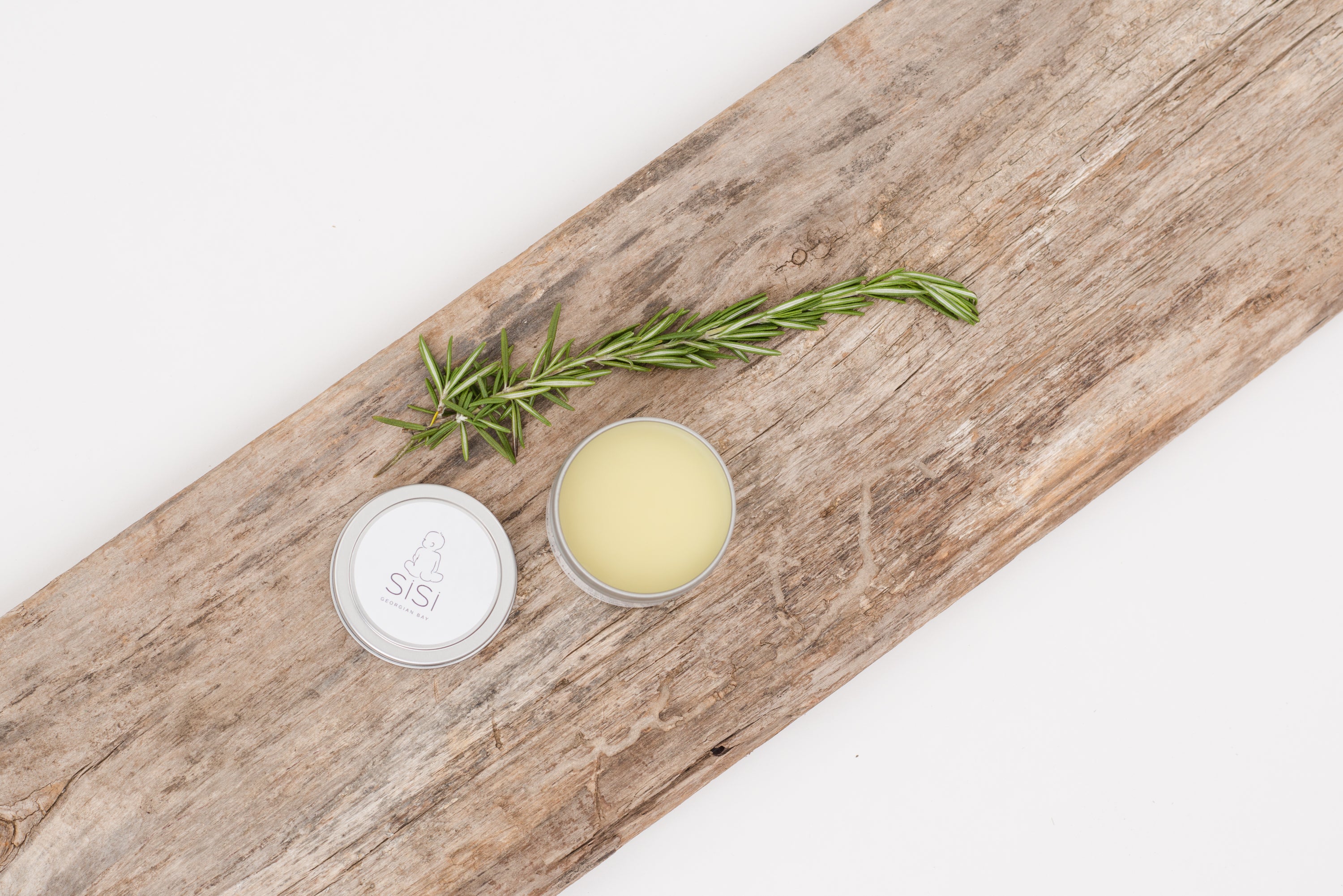 natural skincare products - a photo of a piece of rustic but nice looking wood on a diagonal with a creamy white background with a tin of natural baby balm on it with a green leafy sprig laying beside it