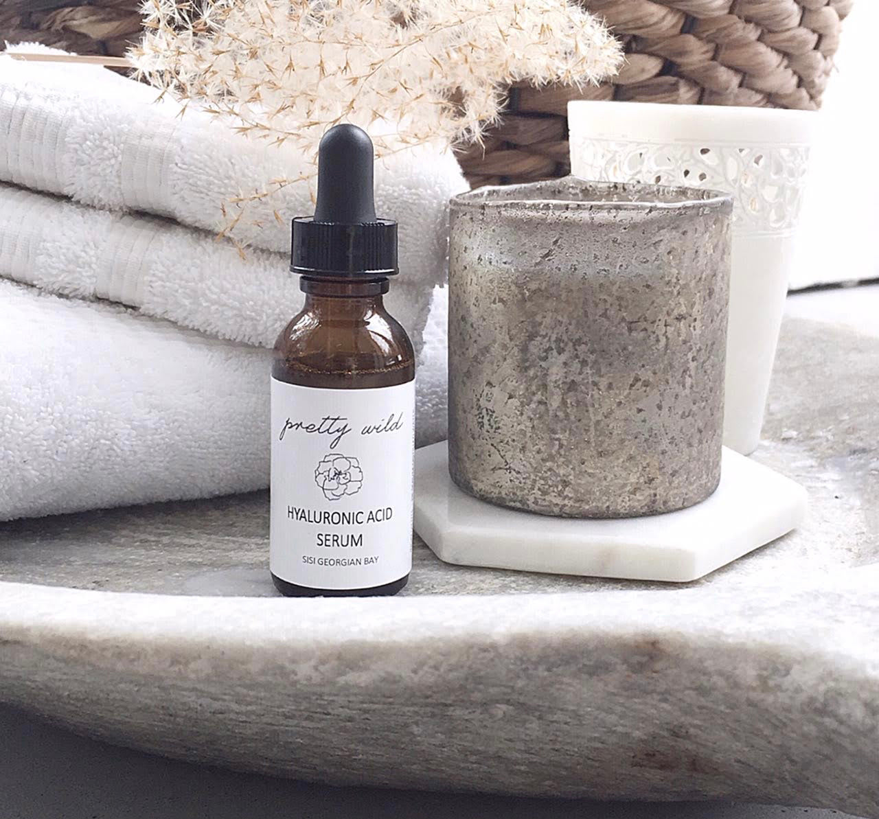 natural skincare products - a photo of the pretty wild hyaluronic acid serum in a 30ml amber glass bottle in front of a beautiful stone tray with fluffly white towels and a wicker basket behind it and a pottery cup beside it