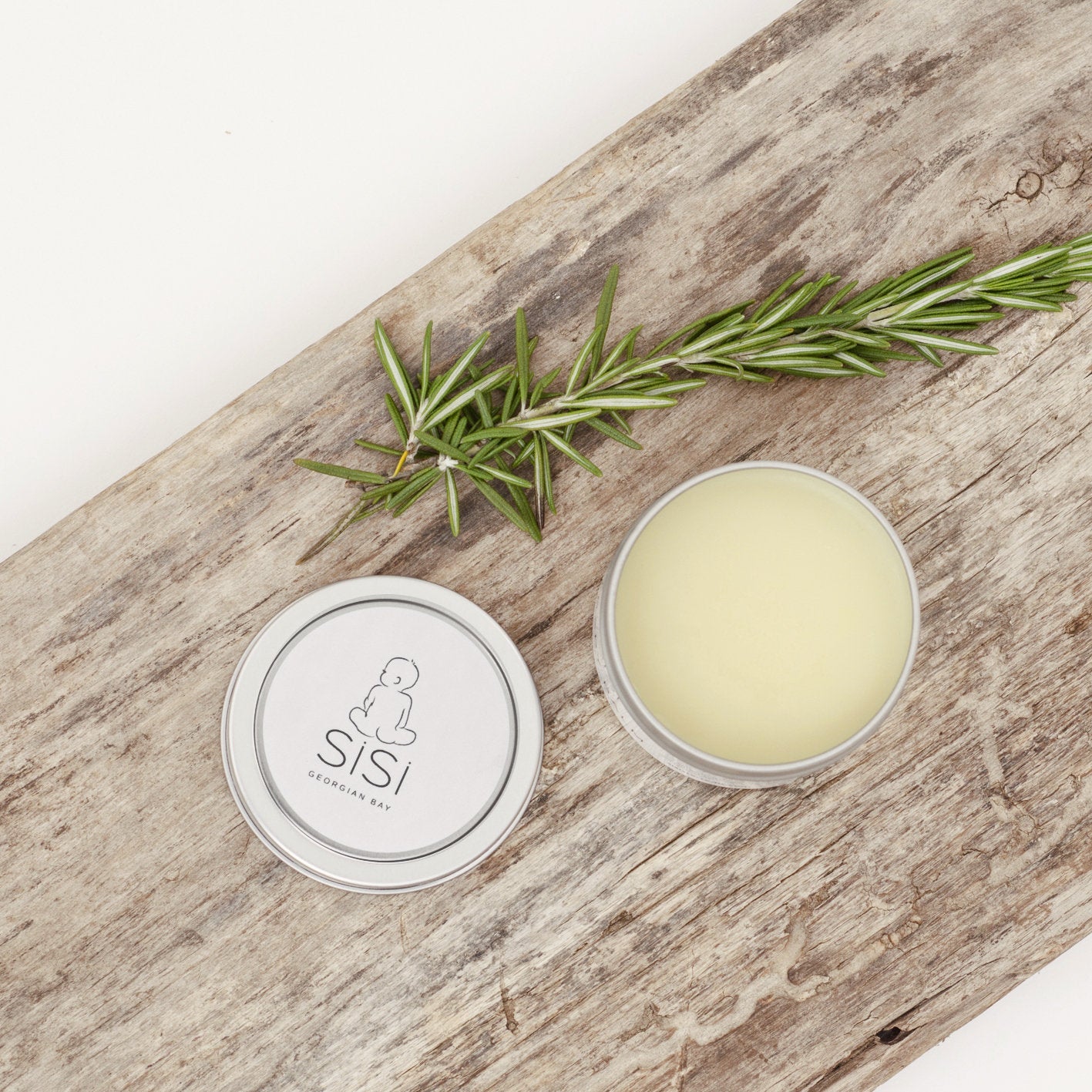 A lovely picture of the baby blam on a rustic wooden piece of wood with the lid off. The balm looks smooth and is pale yellow. The lid is beside the tin with an adorable labe that shows a hand drawn baby and the SiSi Georgian Bay logo