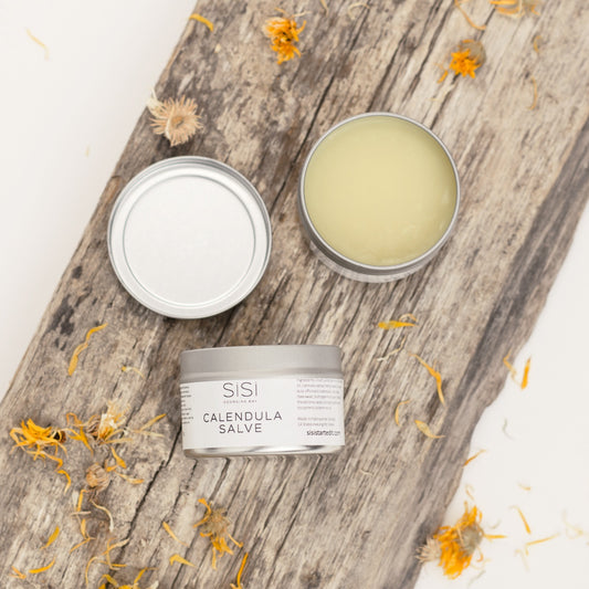 SiSi Georgian Bay Calendula Salve with the lid off on a rustic wooden board with calendula flowers sprinkled around it