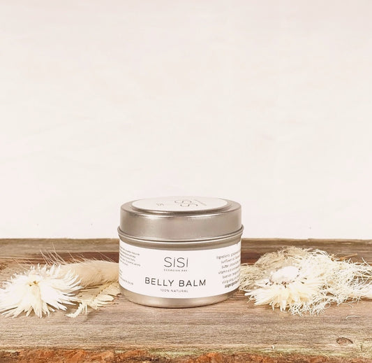 A 4oz tin of Belly Balm on a wooden table top with pretty white flowers sprinkled around it