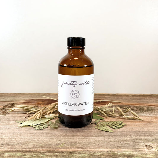 handcrafted small batch natural skincare products - SiSi Georgian Bay "pretty wild" micellar water in an apothecary inspired amber glass bottle on a rustic wooden surface with natural leaves spread out around it