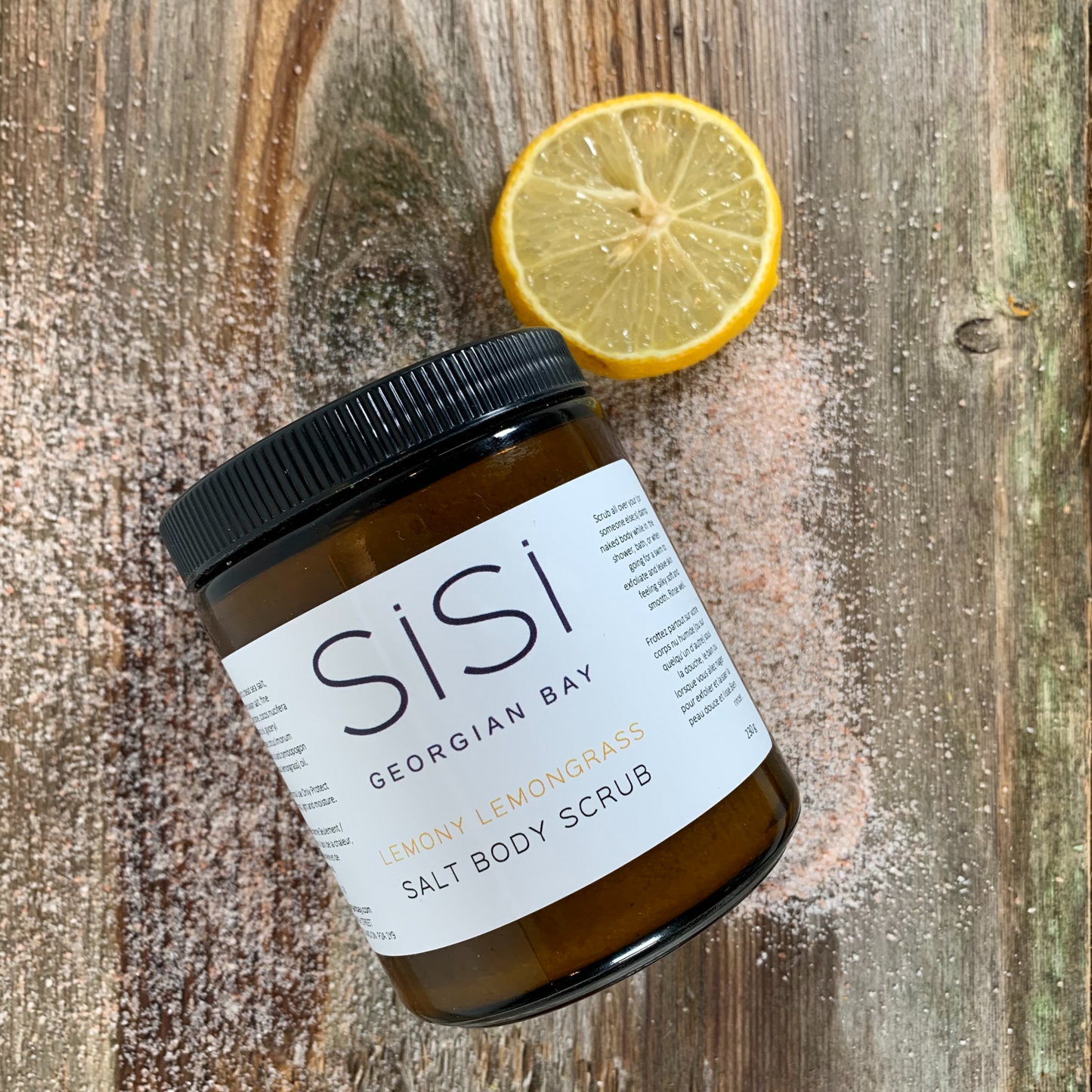 SiSi Georgian Bay Lemony lemongrass salt scrub in an amber glass jar on a rustic wood surface with a lemon slice beside it and salt sprinkled around it