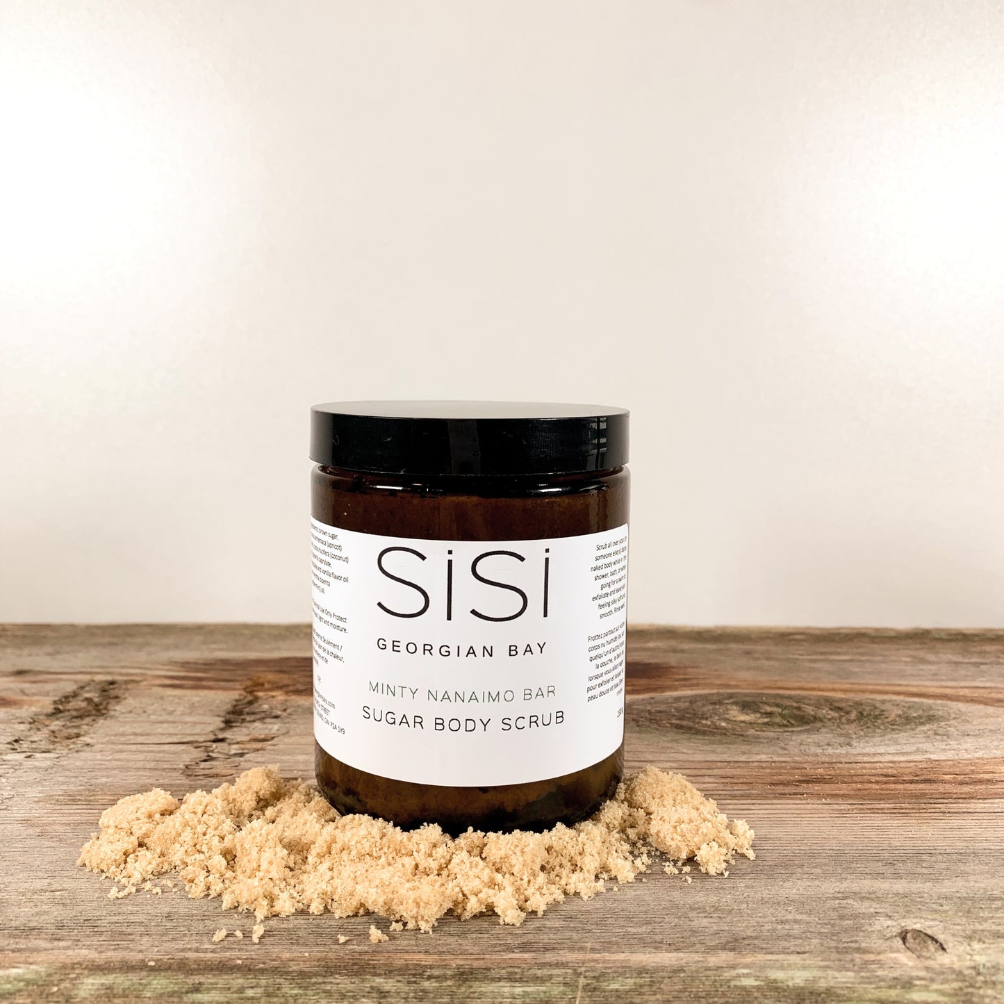 A jar of SiSi Georgian Bay Minty nanaimo bar sugar body scrub on a wooden counter with brown sugar around it