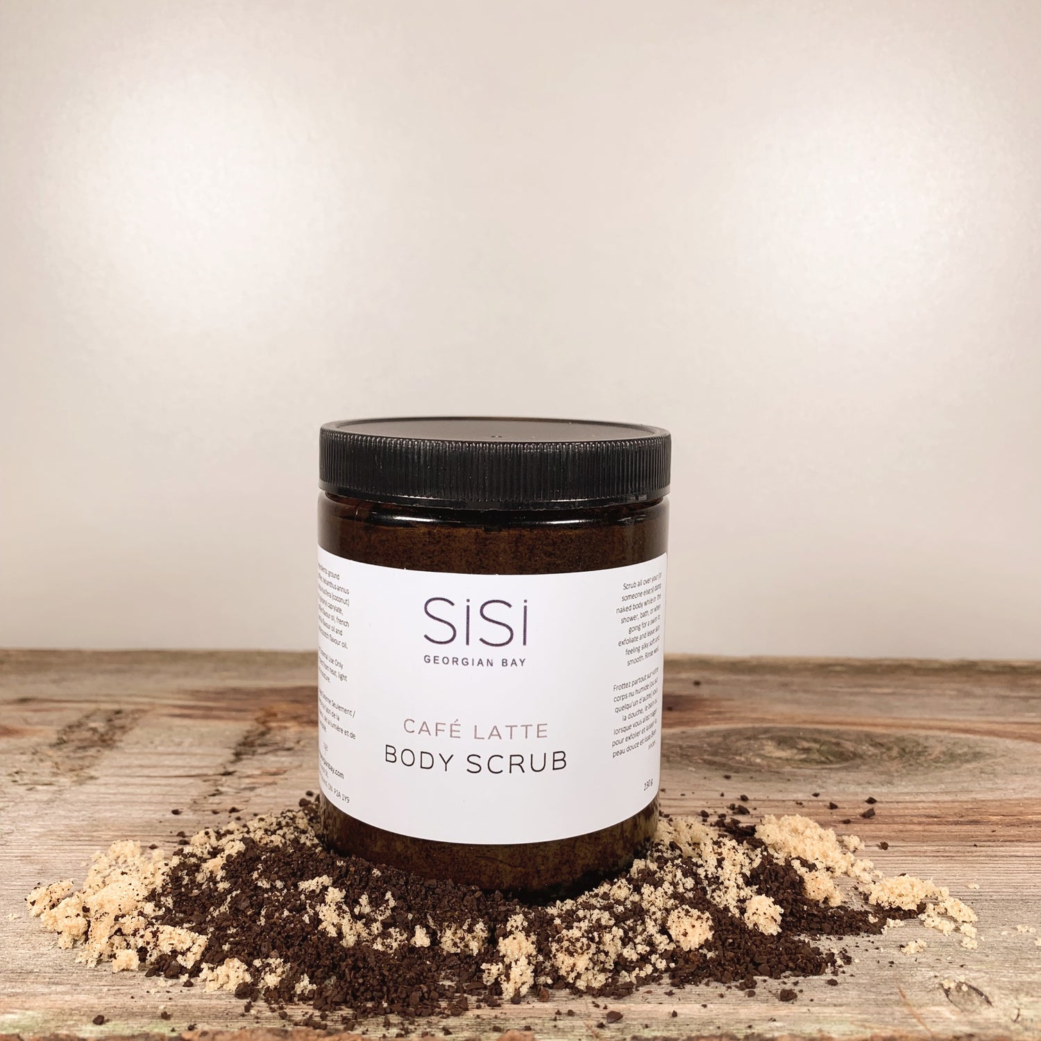 a jar of sisi georgian bay cafe latte coffee body scrub on a wooden counter with coffee grounds and brown sugar sprinkled around it