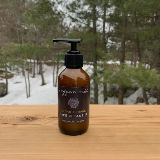 Natural skincare for him. SiSi Georgian Bay 'rugged wild" face cleanser in an amber glass bottle with a pump lid on a wooden table outside in nature with green evergreen trees and snow in the background
