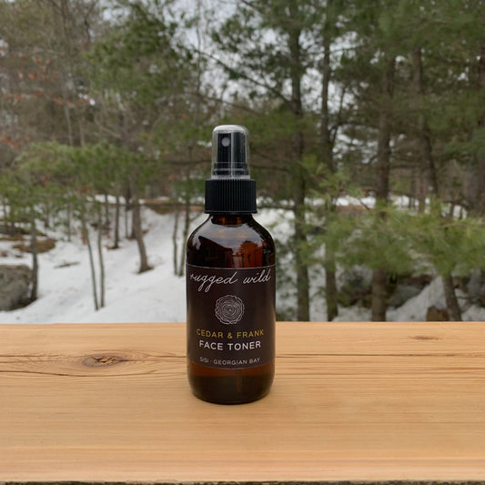 Natural skincare for him. SiSi Georgian Bay 'rugged wild" face toner in an amber glass bottle with a mister lid on a wooden table outside in nature with green evergreen trees and snow in the background