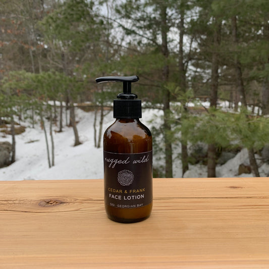 Natural skincare for him. SiSi Georgian Bay 'rugged wild" face lotion in an amber glass bottle with a pump lid on a wooden table outside in nature with green evergreen trees and snow in the background