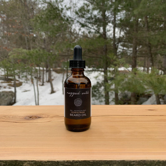 a 60ml amber glass bottle with a glass dropper top with our rugged wild label with a hand drawn slice of wood. The bottle is on cedarwood with a natural background of green trees with a rocks peeking out of a snow covered ground