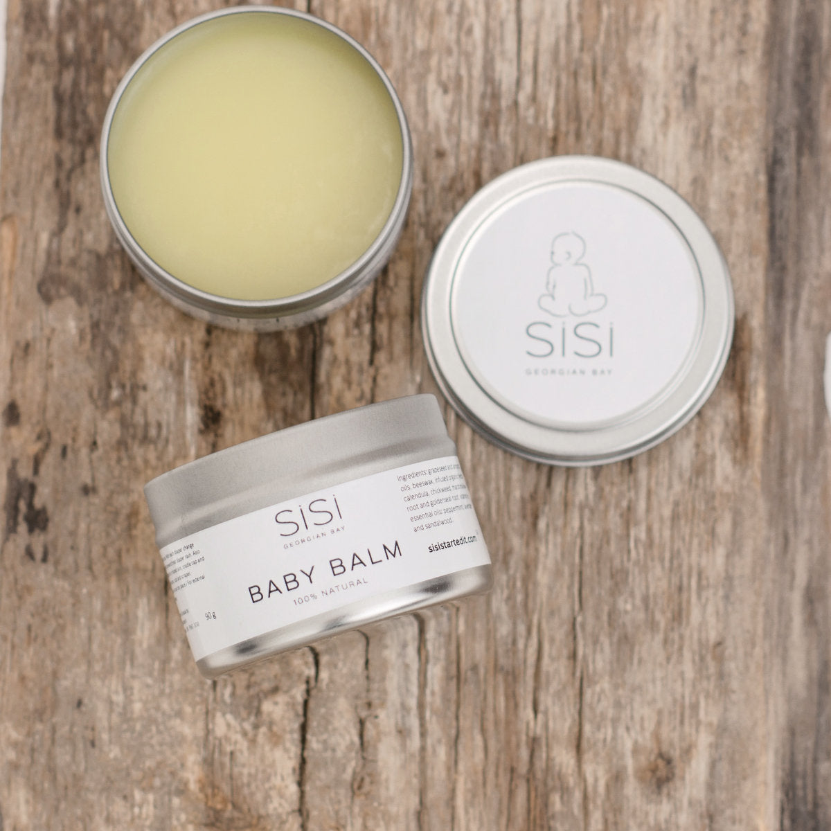 A close up picture of the baby balm with the lid off and laying beside the balm on a rustic piece of wood with another tin of baby balm beside it on it's side