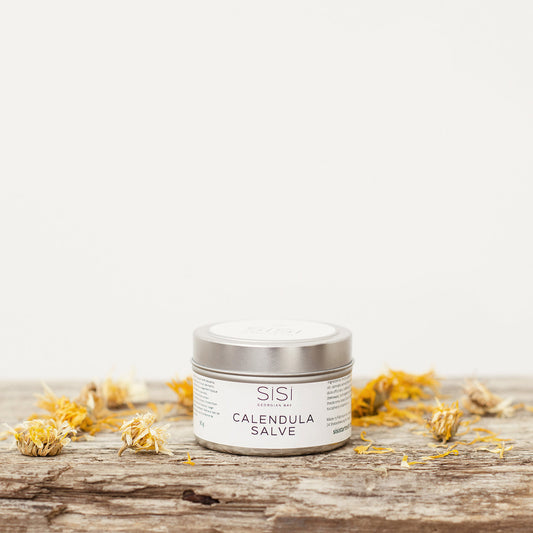 A silver tin of SiSi Georgian Bay Calendula Salve on a rustic wooden surface with calendula flowers sprinkled around it